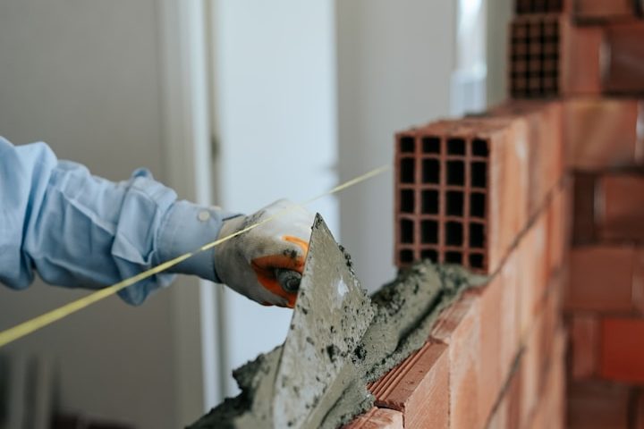 Ein Bauarbeiter vermischt Mörtel und misst mit einem Maßband neben einer Ziegelwand.