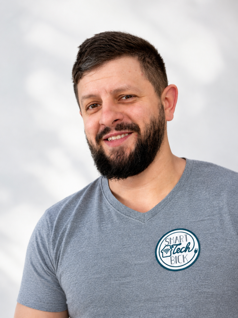 Mann mit Bart, lächelnd, trägt ein graues T-Shirt mit Logo. Heller Hintergrund.