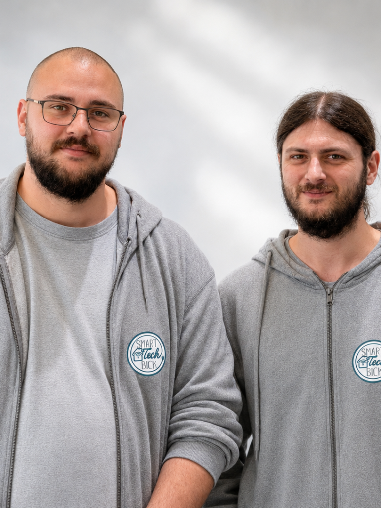 Zwei Männer mit Brillen in grauen Sweatshirts, freundlich lächelnd.